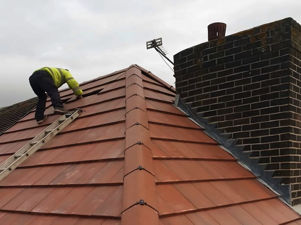Red tile hip with clipped ridge tiles running up to a brick chimney with stepped lead flashing, roofer on the slope