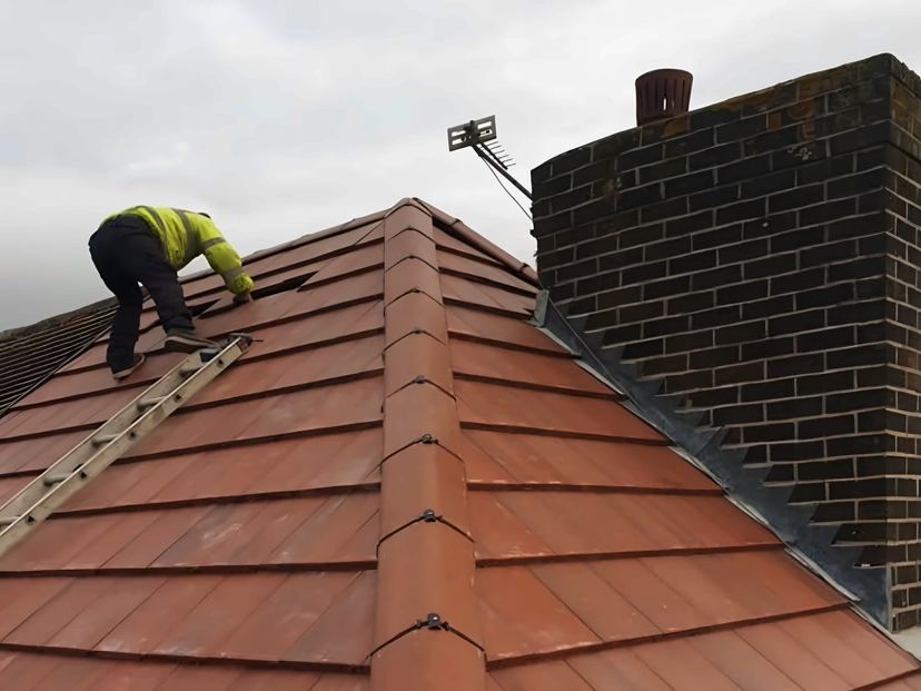 Red tile hip with clipped ridge tiles running up to a brick chimney with stepped lead flashing, roofer on the slope