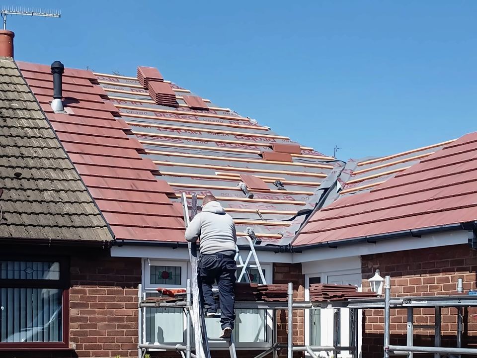 The same bungalow part-way through: membrane and battens on, red tiles loaded out, roofer on the ladder