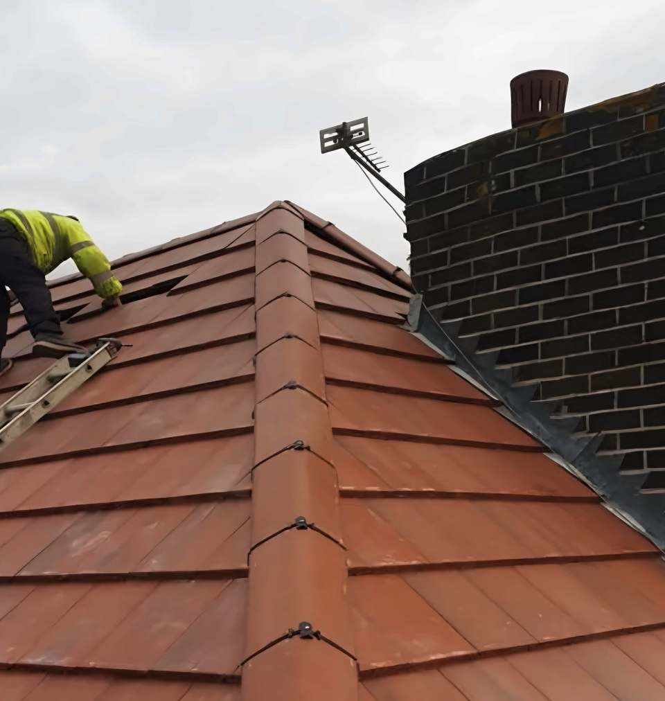 Red tile hip with clipped ridge tiles running up to a brick chimney with stepped lead flashing, roofer on the slope